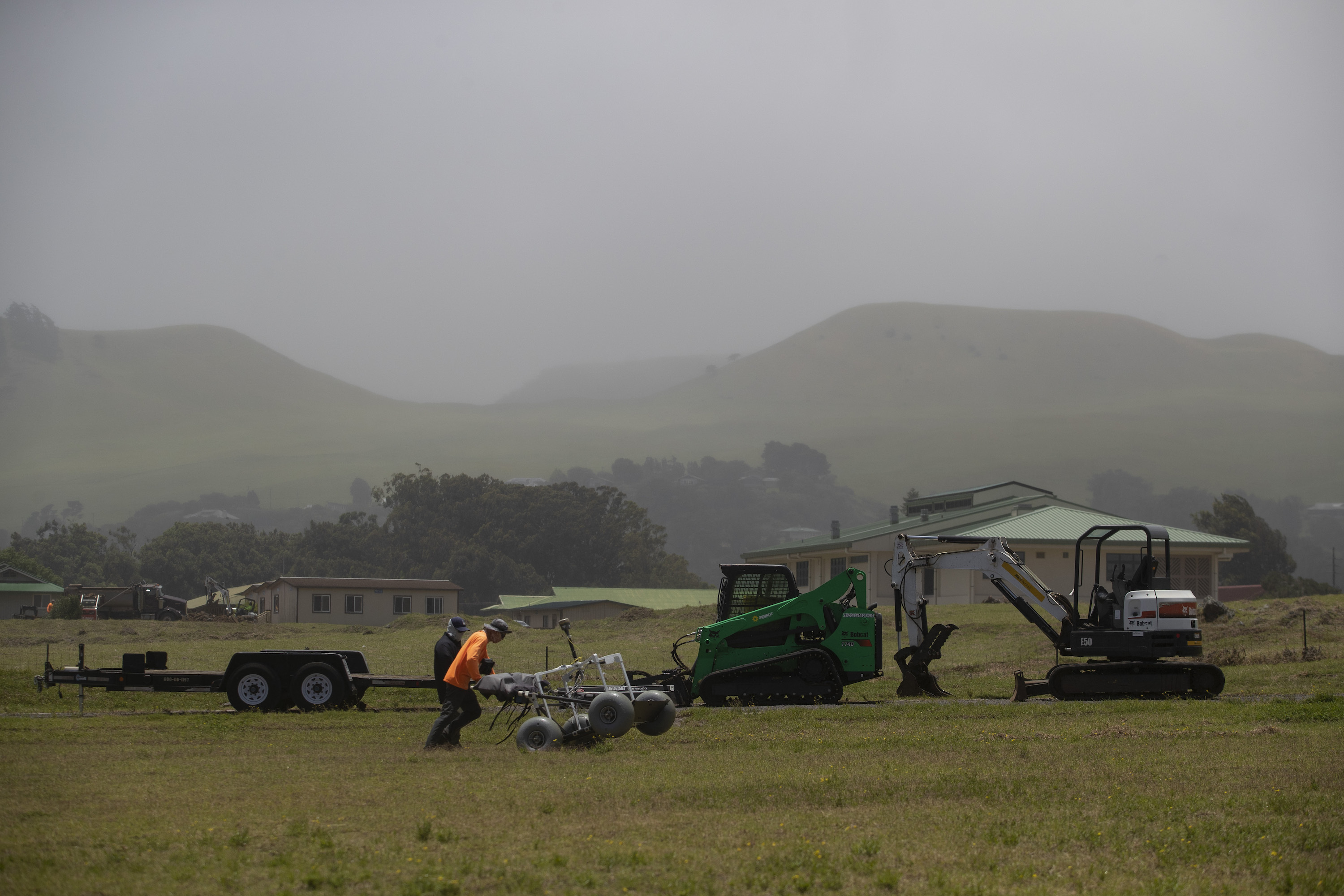 A man in an orange shirt and baseball cap pushes a four-wheeled device with balloon tires across a field.
